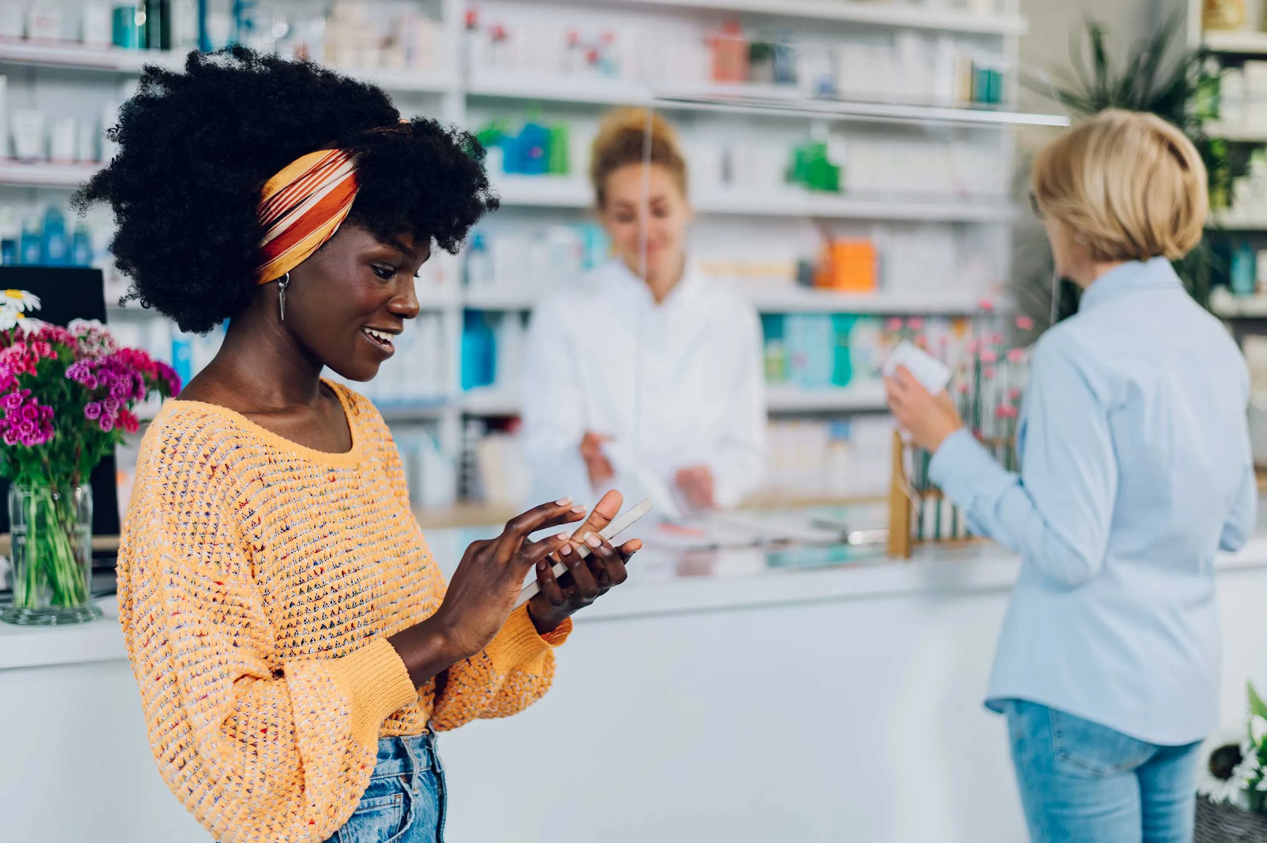 Eine junge Frau nutzt ihr Handy in der Apotheke, während im Hintergrund eine andere Frau von der Apothekerin bediehnt wird